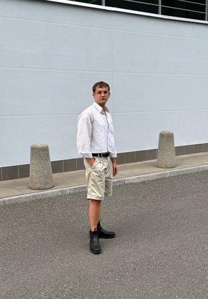 Camisa de botones blanca a rayas verticales, pantalones cortos beige y botas negras hasta el tobillo. Modelo de pie contra una pared gris claro sin decoración.