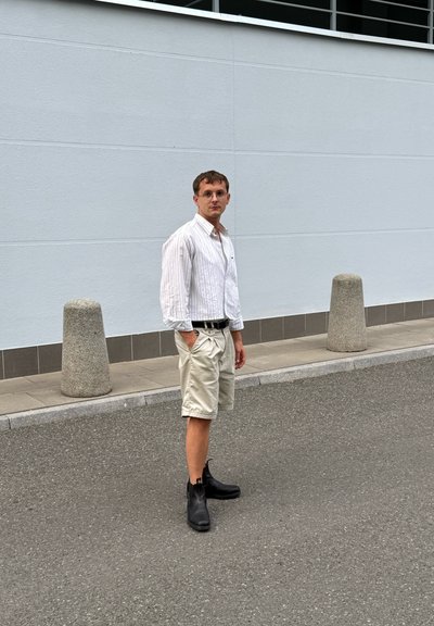 Camisa de botones blanca a rayas verticales, pantalones cortos beige y botas negras hasta el tobillo. Modelo de pie contra una pared gris claro sin decoración.