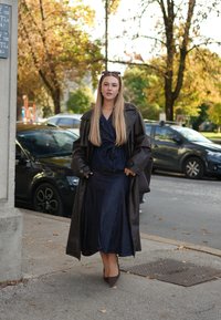 Brązowy skórzany płaszcz trench na ciemnej dżinsowej sukience. Strój ma fason kopertowy, długie rękawy oraz brązowe szpilki z zakończeniem w szpic.