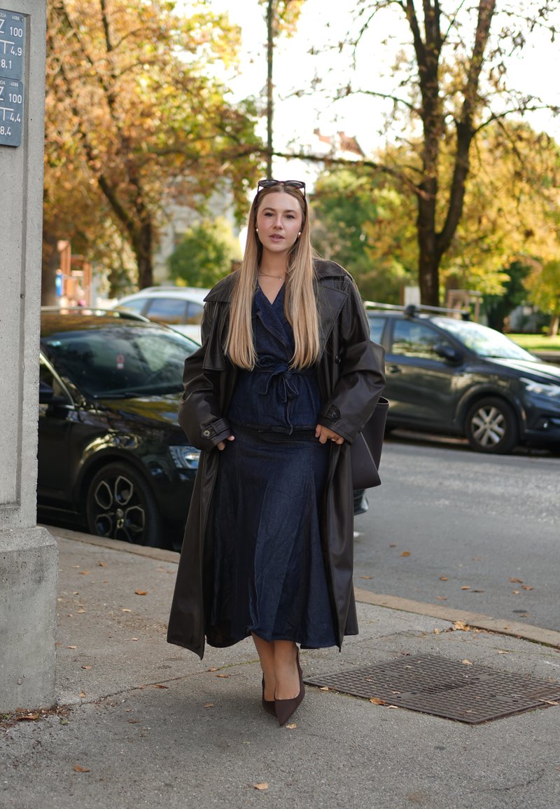 Brauner Leder-Trenchcoat über einem dunklen Denim-Kleid. Das Outfit zeichnet sich durch ein Wickeldesign, lange Ärmel und spitze braune Absatzschuhe aus.