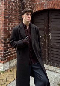 Lang mørk frakke og dyb bordeaux skjorte, kombineret med en sort handske. Står op ad en murstensvæg og trædøre. Fladt fortov.