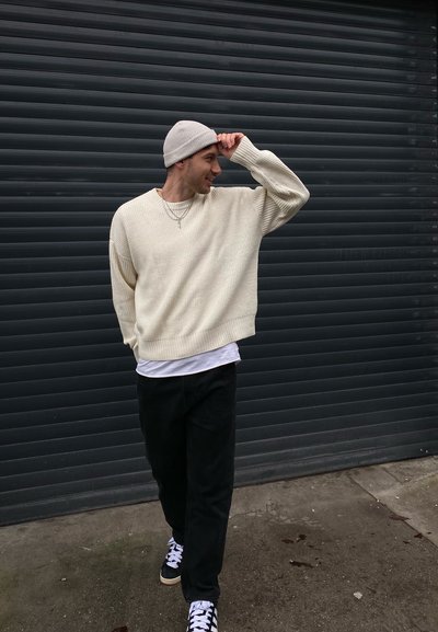 Suéter blanco de canalé, pantalones negros y zapatillas negras con acentos blancos. Gorro beige, con una camiseta blanca debajo.