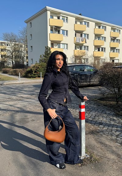 Modelo con una chaqueta de mezclilla oscura ajustada y pantalones vaqueros oscuros holgados, sosteniendo un bolso marrón, de pie al lado de un poste rojo y blanco en una calle.