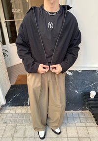 Sort lynlåstrøje over en sort t-shirt med hvidt logo, sat sammen med beige vidde-bukser og tofargede sko. Sølvkædehalskæde.