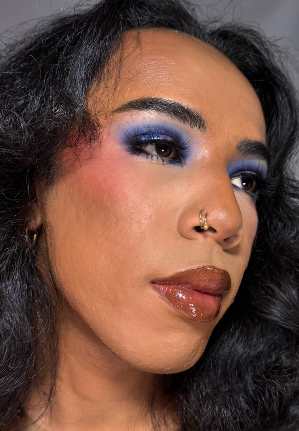 Gros plan sur un visage avec des cheveux noirs ondulés, portant un fard à paupières bleu vif, un blush subtil, des lèvres brunes brillantes et un anneau de nez en or.