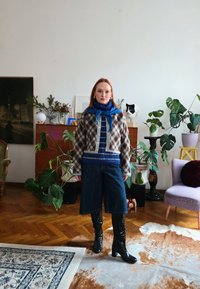 Γυναίκα με ζακέτα με σχέδια, μπλε κασκόλ, ριγέ πουλόβερ, τζιν κολάν και μαύρες μπότες που στέκεται σε σαλόνι γεμάτο φυτά με ξύλινο πάτωμα.
