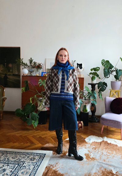 Mujer con cárdigan estampado, bufanda azul, suéter de rayas, pantalones culottes de mezclilla y botas negras, de pie en una sala llena de plantas con suelo de madera.