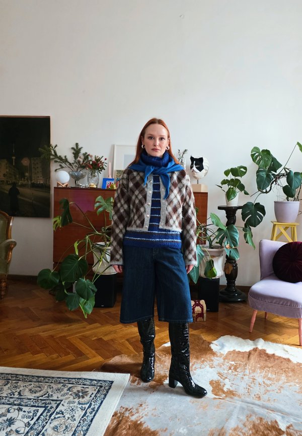 Sieviete ar raibu kardiganu, zilu šalli, svītrainu džemperi, džinsa kulotēm un melniem zābakiem stāv telpā ar daudzām augiem un koka grīdu.