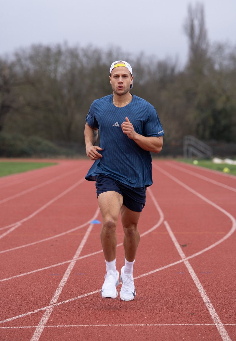 Man in blauwe sportkleding en witte pet die op een buitenbaan met rode ondergrond rent, met bomen op de achtergrond.