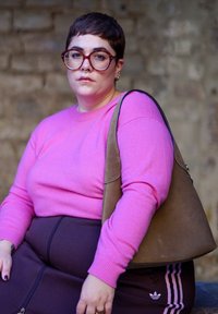 Ροζ πουλόβερ, καφέ φούστα με ψηλή μέση και φερμουάρ, και μια μεγάλη μπεζ τσάντα. Κοντά μαλλιά, στρογγυλά γυαλιά και διακριτική τρύπα στη μύτη. Υφή φόντου.