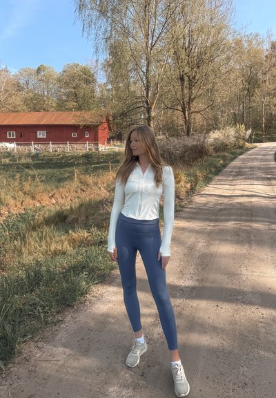 Camisa blanca de manga larga con cierre de cremallera, combinada con leggings azules de cintura alta. Neutros en las zapatillas, ambiente casual al aire libre con vegetación.