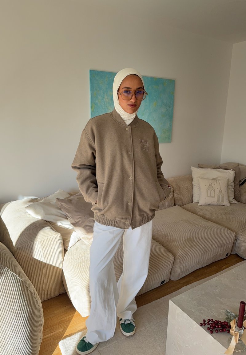 Veste en polaire marron avec fermeture à boutons, ornée d'un patch logo, associée à un pantalon large blanc et des baskets vertes, dans une pièce lumineuse.