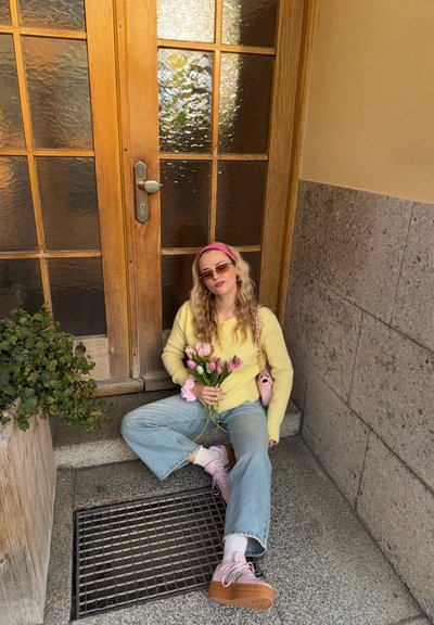Mujer con gafas de sol, suéter amarillo y zapatos rosas sentada en escalones de piedra sosteniendo tulipanes rosas junto a una puerta de madera con paneles de vidrio esmerilado.