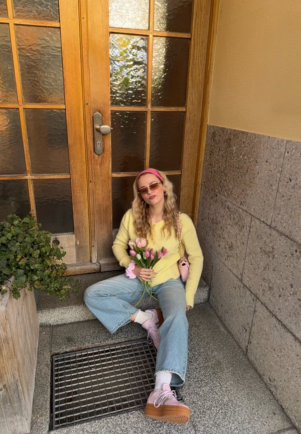 Žena s okuliarmi na slnko, žltým sveterom a ružovými topánkami sedí na kamenných schodoch a drží ružové tulipány pri drevených dverách s matnými sklenenými panelmi.