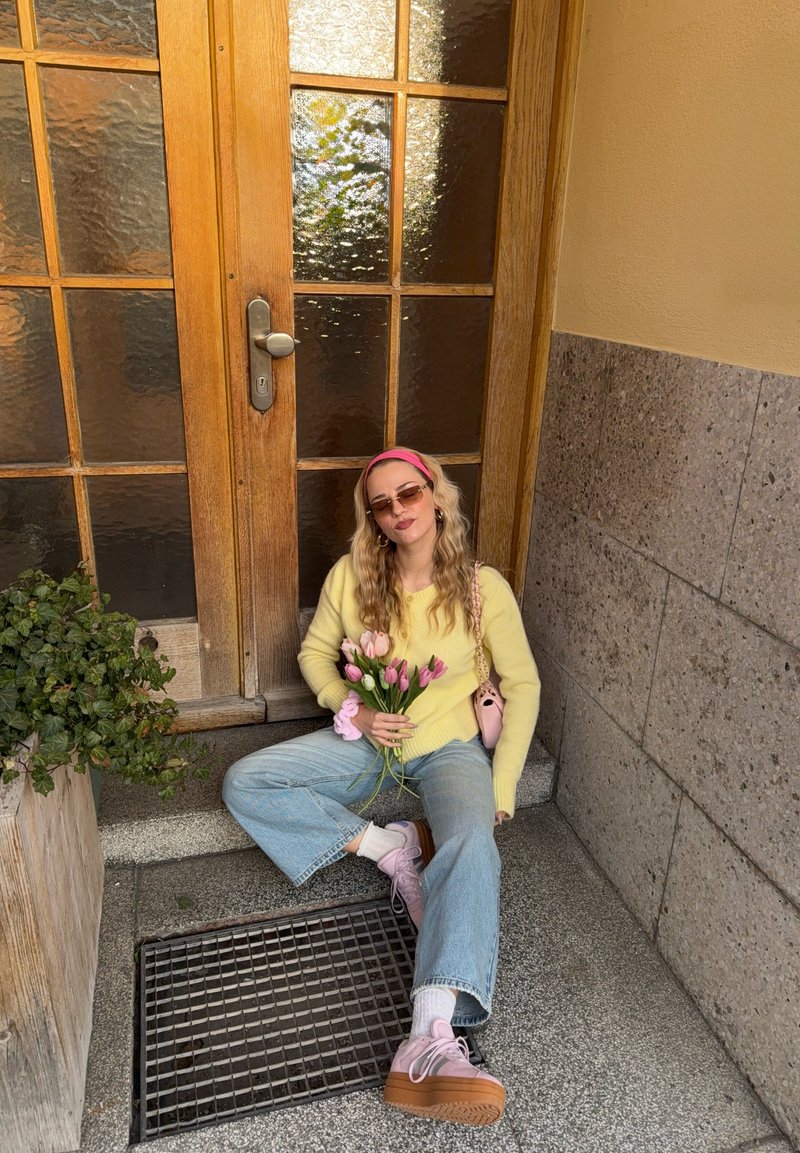 Femme portant des lunettes de soleil, un pull jaune et des chaussures roses, assise sur des marches en pierre, tenant des tulipes roses près d'une porte en bois avec des panneaux en verre dépoli.