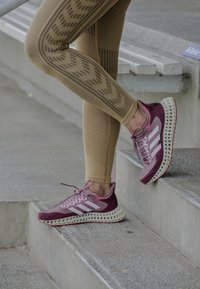 Vinröda träningsskor med vita ränder på sidan, texturerad mesh-överkonstruktion och en distinkt nätverksdesign på sulan.