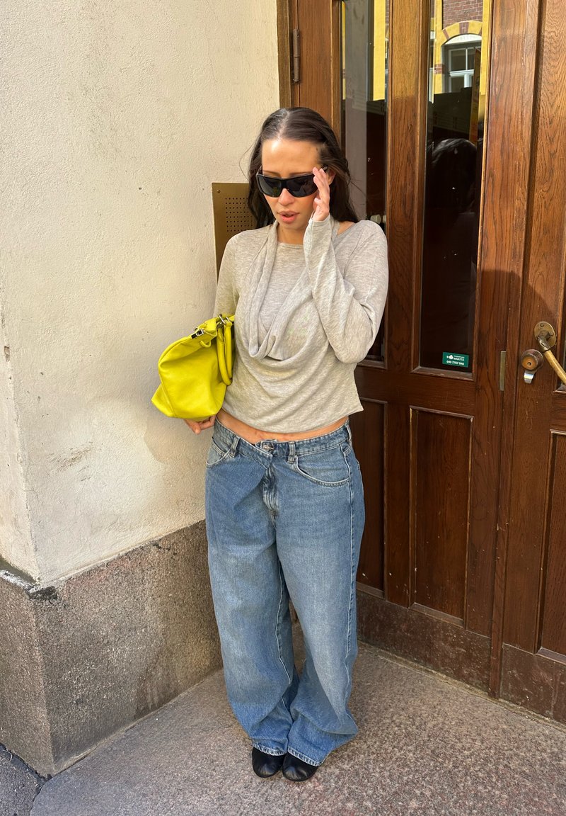 Frau in lockeren Jeans, hellgrauem Langarmshirt und schwarzen Sonnenbrillen hält eine leuchtend gelbe Handtasche an einer braunen Holztür.