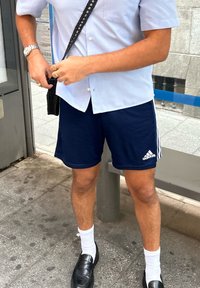 Camisa de botão de manga curta azul-claro e calções de treino azul-marinho com riscas brancas. Bolsa de ombro preta e mocassins pretos brilhantes.