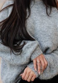 Pull gris en maille texturée avec manches longues ; mains visibles, ornées de bagues et d'ongles clairs, avec des poignets duveteux.
