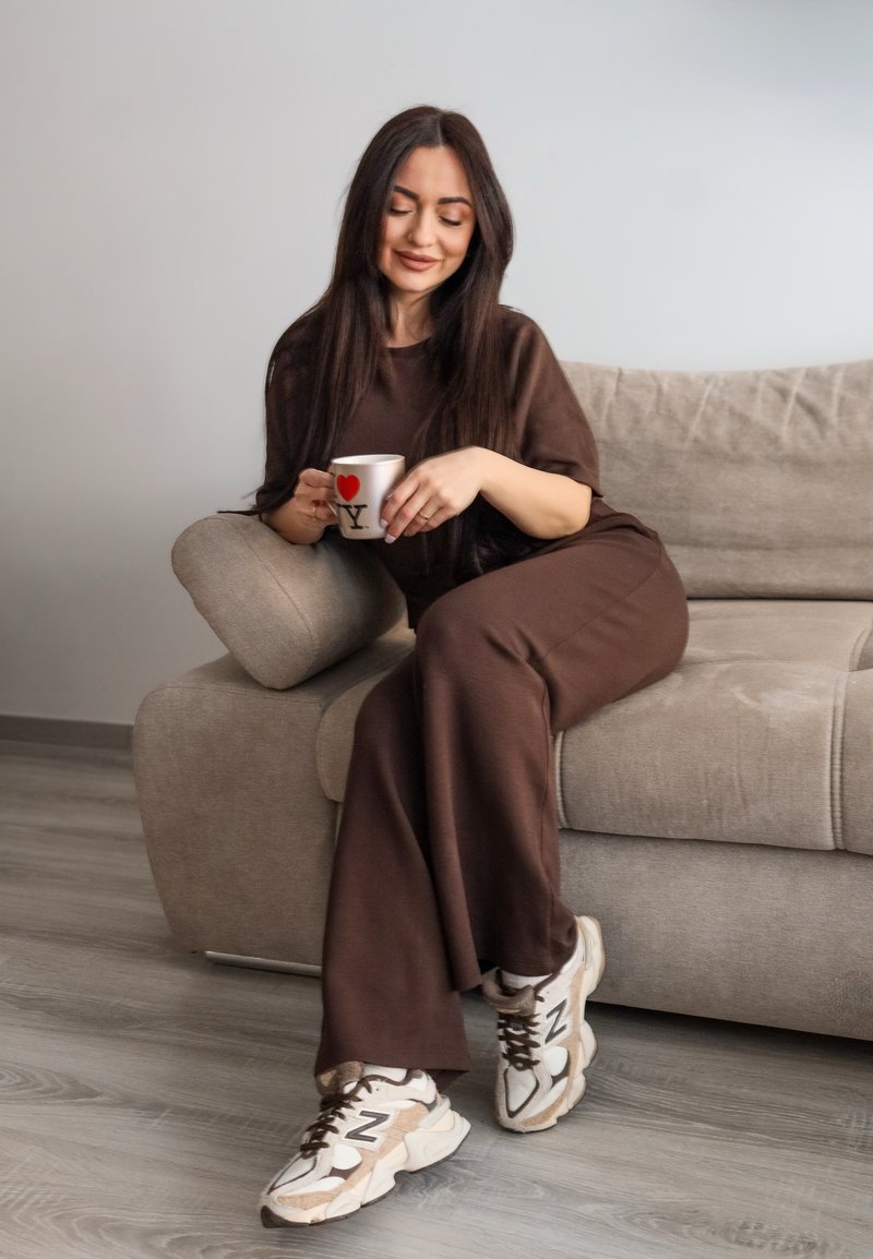 Ruda dviejų dalių laisvo kirpimo apranga, sėdinti ant šviesiai pilkos sofos, laikanti baltą puodelį su raudona širdimi ir raide „Y“, avinti kreminės ir smėlio spalvos sportbačiai.