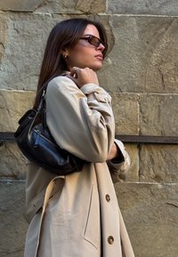 Kvinne i beige trenchcoat og solbriller som bærer en svart lær skulderveske mot en steinvegg.