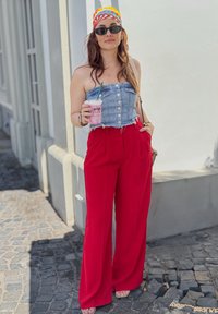 Žena nosi traper bezrameni top s dugmadi, crvene široke hlače, crne sunčane naočale i šareni marama na glavi, držeći piće.