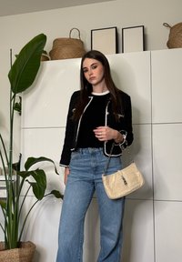 Jeune femme aux longs cheveux bruns portant une veste noire et un jean bleu, tenant un sac à main beige, debout à côté d'une grande plante verte dans une pièce moderne.