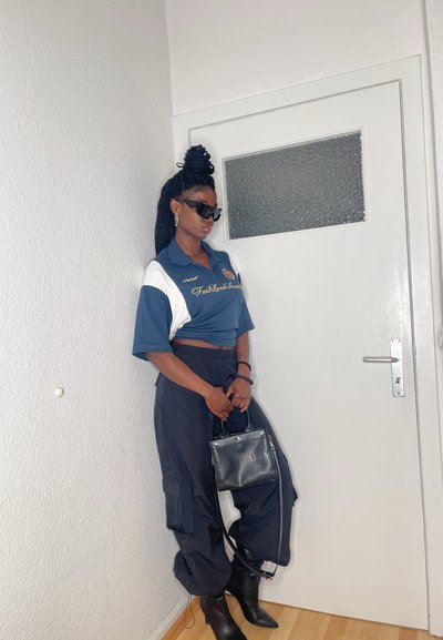 Polo recortado azul marino y blanco con bordado dorado, pantalones cargo negros, bolso negro y botas negras de punta; la modelo lleva gafas de sol.