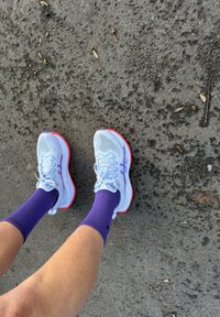 Chaussures de course bleu clair avec semelles roses, dotées d'une tige en mesh et de lacets, accompagnées de chaussettes de sport violettes, sur une surface grise texturée.