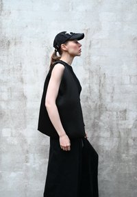 Top senza maniche nero con texture e design asimmetrico, abbinato a pantaloni neri larghi. Gli accessori includono un cappellino nero con un dettaglio grafico.