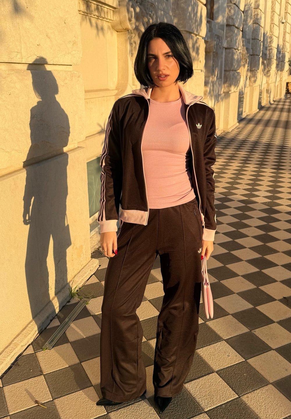 Femme aux cheveux courts noirs portant un survêtement noir et rose avec un haut rose, debout sur un trottoir à damier avec une ombre sur un mur en pierre.