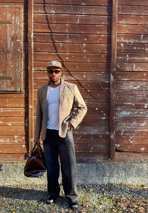 Hombre con blazer beige, gorra gris, gafas de sol, camisa blanca, vaqueros negros y zapatos negros, de pie junto a una pared de madera envejecida con un bolso de cuero marrón.