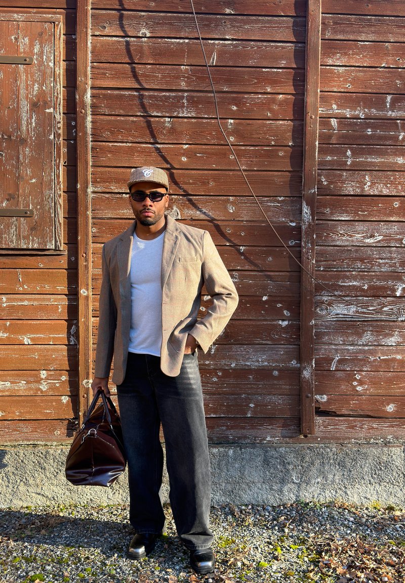 Homme portant un blazer beige, une casquette grise, des lunettes de soleil, une chemise blanche, un jean noir et des chaussures noires, debout avec un sac en cuir marron près d'un mur en bois patiné.