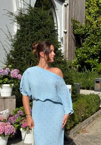 Vestido pregueado azul claro com um design à mostra de ombros, apresentando detalhes prateados e uma textura fluida. Colocado em meio a vegetação e flores.