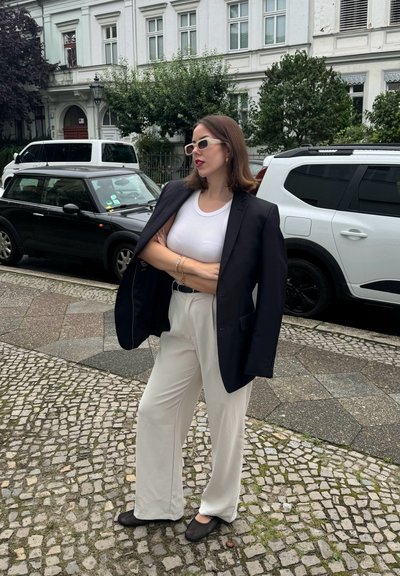 Blazer negro sobre una blusa blanca, combinado con pantalones blancos holgados y bailarinas de malla negra. El fondo presenta coches y una calle de adoquines.