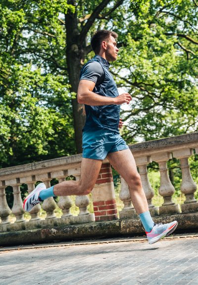 Corredor atlético con un chaleco azul marino y pantalones cortos turquesa, que lleva zapatillas blancas y rosas con calcetines azules, corriendo por un camino pavimentado al lado de una barandilla de piedra.