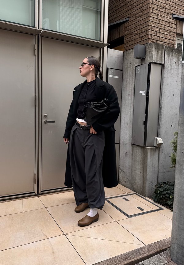 Black coat over a dark shirt, loose grey trousers, and brown shoes with white socks; holding a black bag; standing near metal doors.