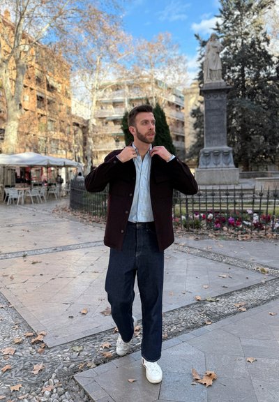 Hombre vestido con una chaqueta abotonada marrón oscuro, una camisa de rayas claras y jeans oscuros holgados, complementados con zapatillas blancas en un entorno urbano.