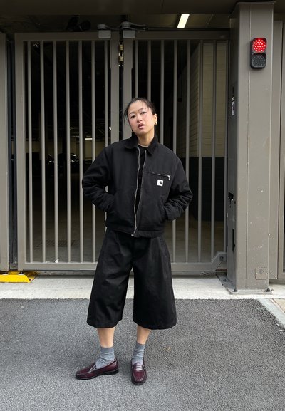 Chaqueta negra con bolsillo con cremallera, pantalones negros amplios y recortados, calcetines grises y mocasines burdeos. De pie frente a un área cerrada.