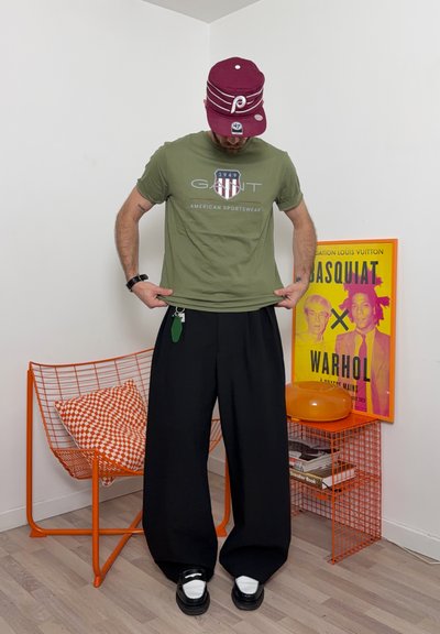 Hombre con camiseta verde oliva de GANT y gorra burdeos, de pie junto a una silla de alambre naranja y un colorido póster de Basquiat Warhol en la esquina de la habitación.