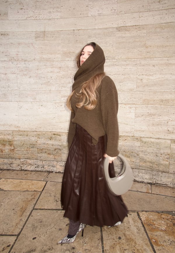 Donna indossa un maglione marrone con cappuccio lavorato a maglia, una gonna plissettata in pelle marrone scuro, tacchi in pelle di serpente e tiene in mano una borsa a mezzaluna di colore grigio chiaro.