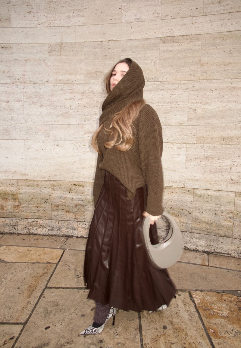 Donna indossa un maglione marrone con cappuccio lavorato a maglia, una gonna plissettata in pelle marrone scuro, tacchi in pelle di serpente e tiene in mano una borsa a mezzaluna di colore grigio chiaro.