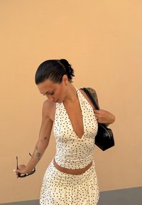 Witte haltertop en bijpassende rok met zwarte stippen, textuurstof, gecombineerd met een zwarte handtas. Achtergrond is een licht perzikkleurige muur.