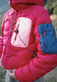Osoba oblečená v červené péřové bundě s růžovou náprsní kapsou a modrou zipovou taštičkou na levém rukávu, drží venku lano.