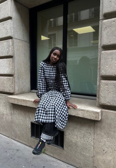 Vestido a cuadros en blanco y negro con mangas abullonadas, sentado en un soporte de piedra, combinado con jeans oscuros y zapatillas negras. Fondo de ventanales.
