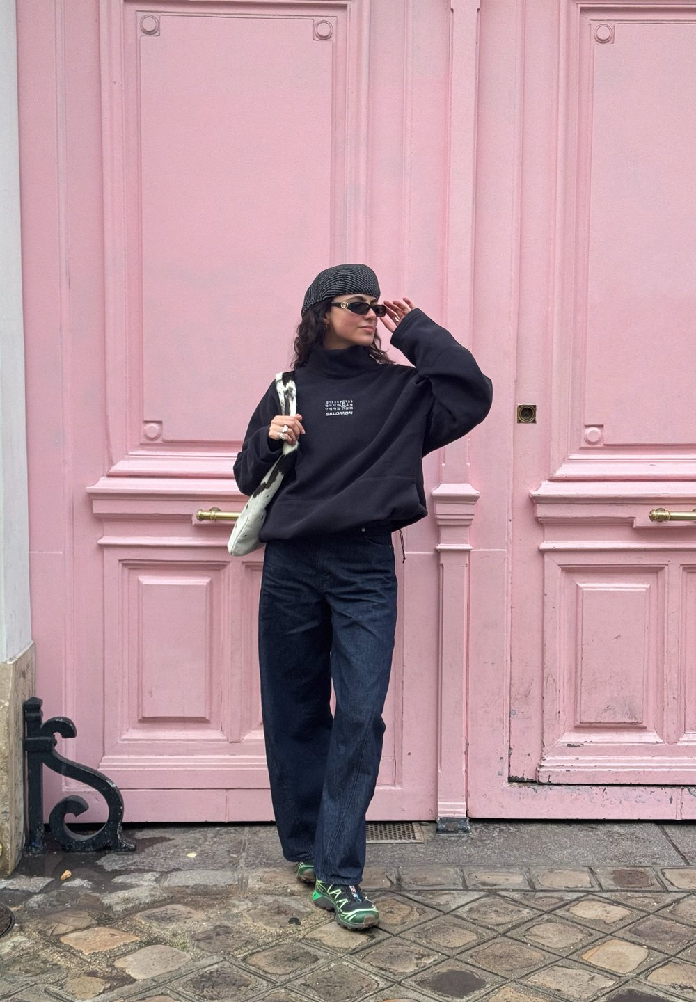 Femme portant un pull noir, un pantalon bleu marine, des baskets vertes, des lunettes de soleil et un chapeau à rayures, debout devant une grande porte en bois rose.
