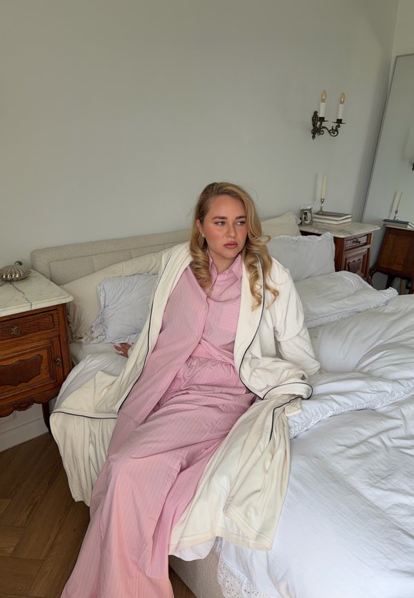 Žena so svetlými vlasmi v ružovom pyžame a bielom župane sediaca na posteli v spálni s dreveným nábytkom a svietiacimi nástennými lampami.