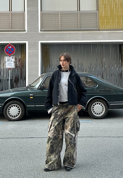 Chaqueta corta negra sobre un suéter gris, combinada con pantalones cargo de camuflaje. Auriculares visibles; de pie frente a un coche verde oscuro.