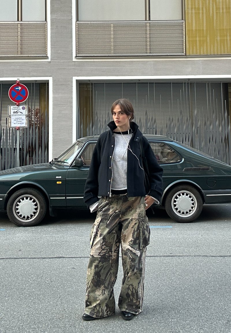 Nero giubbotto corto sopra un maglione grigio, abbinato a pantaloni cargo mimetici. Auricolari visibili; in piedi di fronte a un'auto verde scuro.