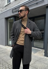 Abrigo de lana gris sobre un suéter de cuello alto beige, pantalones negros, gafas de sol negras y un bolso bandolera. Tejido texturizado con un corte ajustado.